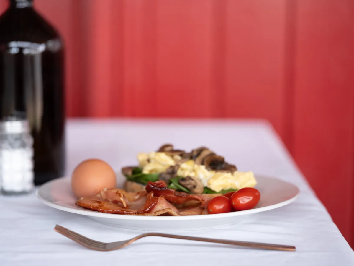 A cooked breakfast plate with eggs, bacon, mushrooms and tomato on a white tablecloth against a red wall.