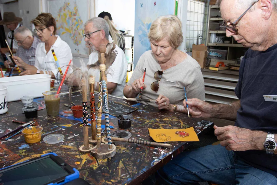 Visitors painting message sticks at a paint-splattered workshop table.