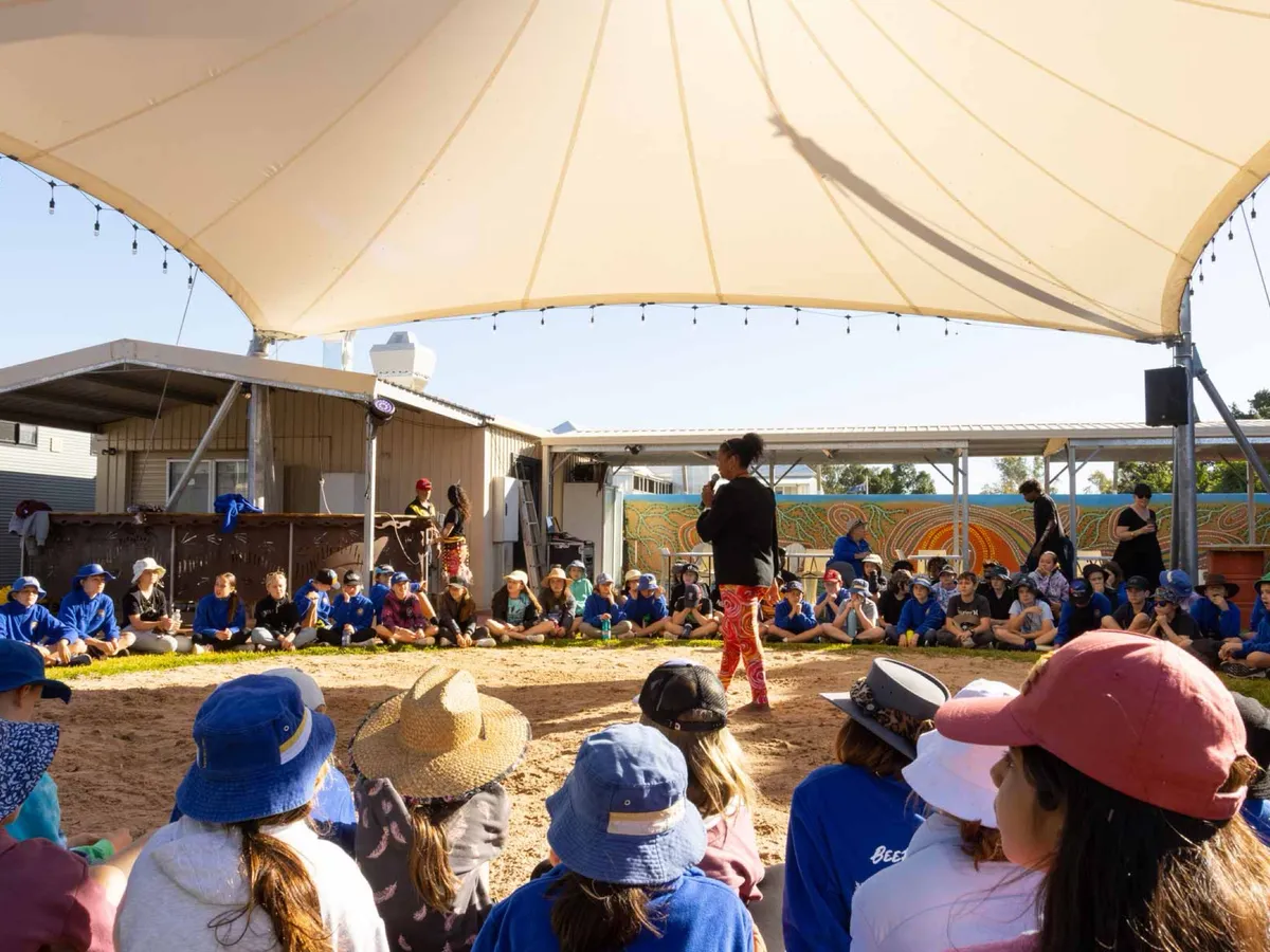 School students in blue uniforms seated in a circle on the Corroboree grounds, listening to a guide standing in the centre.