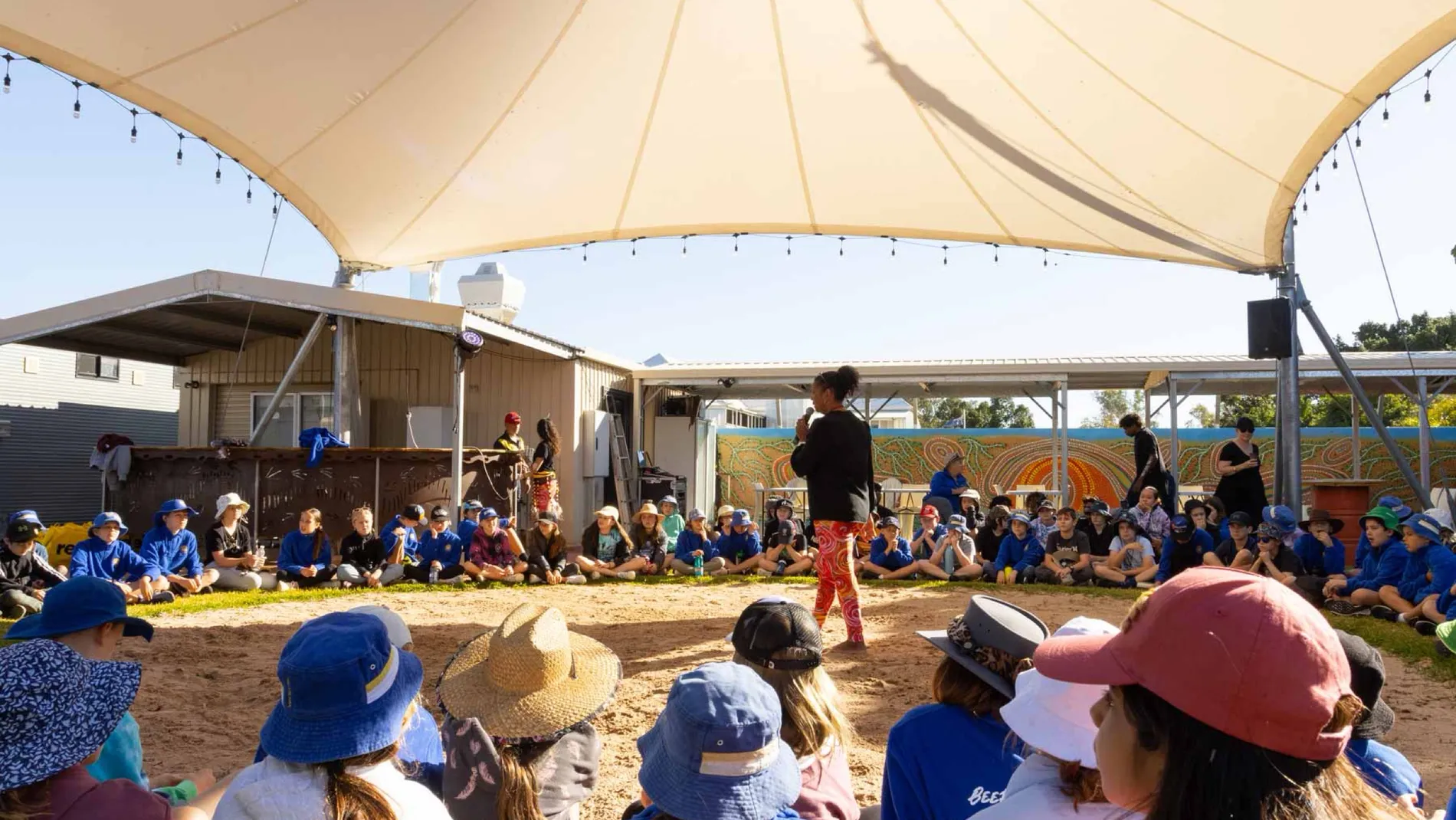 School students in blue uniforms seated in a circle on the Corroboree grounds, listening to a guide standing in the centre.