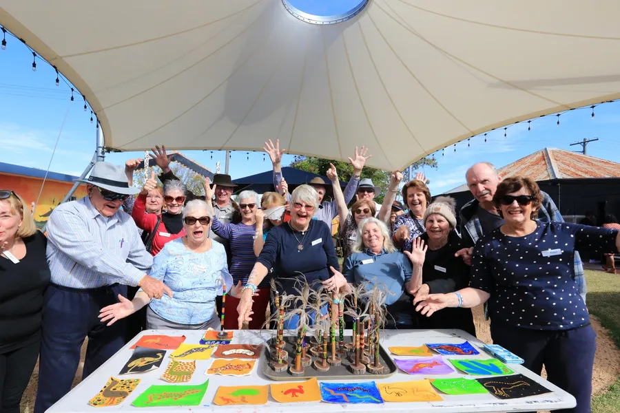 A tour group under the shade structure at the Desert Dreaming Centre, arms raised, with painted artworks and message sticks on the table in front of them.